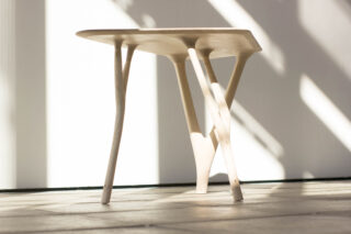 Ein heller Holztisch mit drei einzigartig geformten Beinen steht auf einem gefliesten Boden und wird von der Seite durch natürliches Licht beleuchtet.