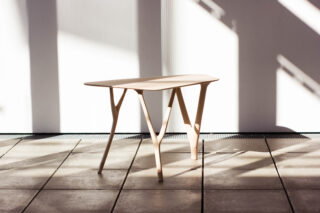 Ein heller Holztisch mit vier dünnen Beinen steht auf einem gefliesten Boden und wird vom Sonnenlicht beleuchtet, das durch ein Fenster strömt und Schatten auf Wand und Boden wirft.