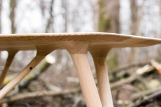 Nahaufnahme eines Holztisches mit glatter Tischplatte und der Verbindung, wo die Beine auf die Unterseite treffen, vor einem unscharfen Waldhintergrund.