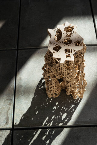 Eine komplexe geometrische Holzskulptur wirft einen detaillierten Schatten auf einen gefliesten Boden, der vom Sonnenlicht beleuchtet wird.