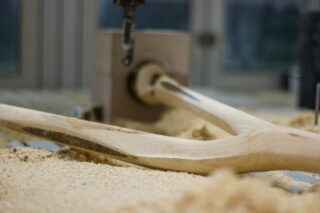 Ein teilweise geschnitzter Baseballschläger aus Holz wird von einer Maschine geformt, wobei Holzspäne im Arbeitsbereich verstreut sind.