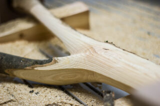 Eine Nahaufnahme eines teilweise geschnitzten Holzobjekts auf einem mit Holzspänen bedeckten Tisch, wobei Holzmaserung und Schnitzspuren sichtbar sind.