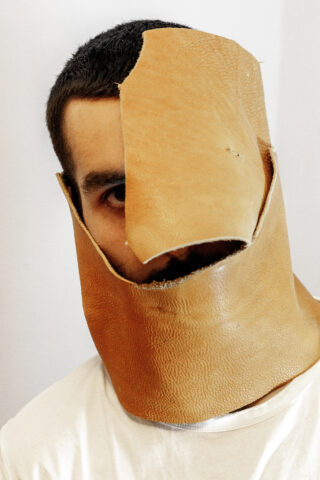 Eine Person, die ein großes, unregelmäßig geformtes Stück hellbraunes Leder trägt, das Teile ihres Gesichts und Halses bedeckt, wobei nur ein Auge und ein Teil ihres Mundes sichtbar sind.