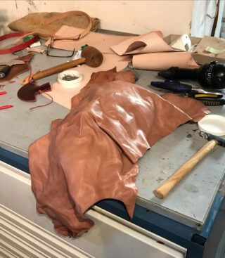 Ein Stück braunes Leder liegt auf einer unordentlichen Werkbank, überall herumliegen verschiedene Werkzeuge und Materialien zur Lederverarbeitung.
