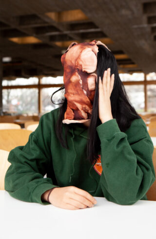 Eine Person in einem grünen Sweatshirt sitzt an einem Tisch. Ihr Gesicht ist mit einer realistischen Maske bedeckt, die rohem Fleisch ähnelt. Sie stützt ihren Kopf auf eine Hand.