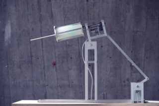 Ein mechanisches Metallgerät mit einem zylindrischen Bauteil und einem hervorstehenden Holzstab, montiert auf einem vertikalen Ständer vor dem Hintergrund einer Betonwand.