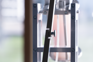 Eine Nahaufnahme eines transparenten zylindrischen Behälters mit Messmarkierungen, teilweise gefüllt mit einer hellrosa Flüssigkeit, montiert auf einem Metallrahmen im Innenbereich.