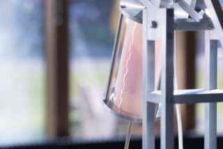 Eine Nahaufnahme eines transparenten zylindrischen Behälters mit Messmarkierungen, teilweise gefüllt mit einer hellrosa Flüssigkeit, montiert auf einem Metallrahmen im Innenbereich.