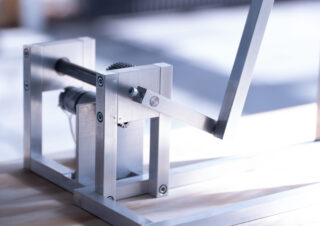 Eine Nahaufnahme eines mechanischen Metallgeräts mit Zahnrädern, Stangen und einem Hebel, das in einer gut beleuchteten Umgebung auf einer Holzoberfläche montiert ist.