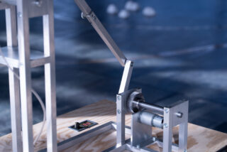 Eine Nahaufnahme eines mechanischen Geräts mit Metallstäben, Zahnrädern und einer kleinen Digitalanzeige, montiert auf einem Holzsockel.