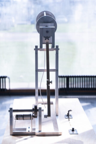 Ein mechanisches Metallgerät mit Zahnrädern, Hebeln und einer vertikalen Stange, auf einem Holzsockel montiert, im Innenbereich mit einem Fenster im Hintergrund aufgestellt.