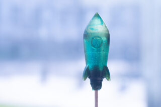 Ein grüner, raketenförmiger Lutscher am Stiel wird aufrecht vor einem unscharfen, hellen Hintergrund gehalten.