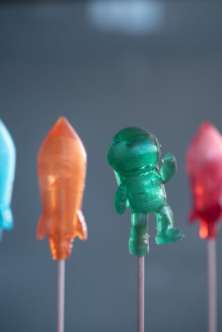 Nahaufnahme von bunten Lutschern in Form einer Rakete und eines Astronauten vor einem unscharfen blauen Hintergrund.