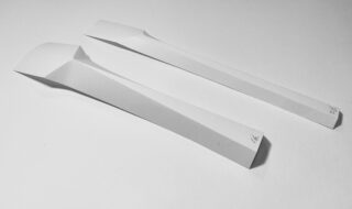 Zwei weiße Papierstreifen, jeweils der Länge nach gefaltet und mit spitzen Enden versehen, werden nebeneinander auf eine weiße Fläche gelegt; an einem Ende jedes Streifens sind kleine Zahlen geschrieben.