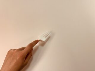 Eine Hand berührt den Griff einer weißen Plastikgabel, die auf einer schlichten, hellen Oberfläche liegt.