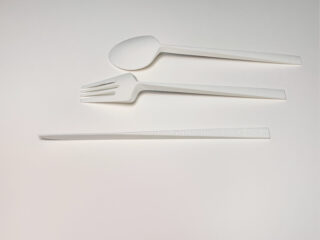 Ein weißer Plastiklöffel, eine Gabel und ein Messer sind parallel zueinander auf einer schlichten weißen Oberfläche angeordnet.