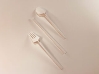 Eine Gabel, ein Löffel und ein Essstäbchen aus weißem Plastik sind parallel auf einer hellbeigen Oberfläche angeordnet.