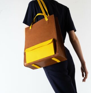 Eine Person in dunklem Outfit trägt vor einem schlichten hellen Hintergrund eine große braun-gelbe Einkaufstasche über der Schulter.