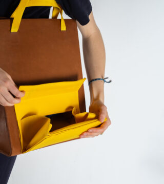 Eine Person öffnet eine braune Ledertasche mit leuchtend gelben Innenfächern vor einem schlichten weißen Hintergrund.