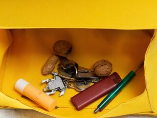 Offene gelbe Tasche mit Lippenbalsam, einem Schlüsselanhänger mit Schlüsseln, einem Feuerzeug, einem grünen Stift, zwei Walnüssen und einer Erdnuss.