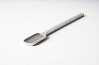 Ein Metallspatel mit rechteckigem Kopf und langem Griff liegt auf einer schlichten weißen Oberfläche.