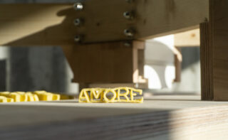 Ein Stück Pasta in der Form von „AMORE“ liegt auf einer Holzoberfläche, im Hintergrund ist ein Teil einer Holzkonstruktion zu sehen.