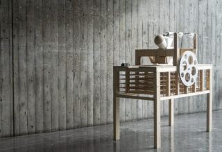 Ein mechanisches Holzgerät mit Zahnrädern und einer kugelförmigen Komponente wird auf einem reflektierenden Boden vor einer strukturierten Betonwand platziert.