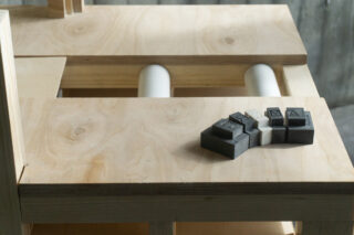 Ein Satz schwarzer und grauer 3D-Puzzleteile liegt auf einer hellen Holzoberfläche mit weiteren Holz- und zylindrischen Komponenten im Hintergrund.