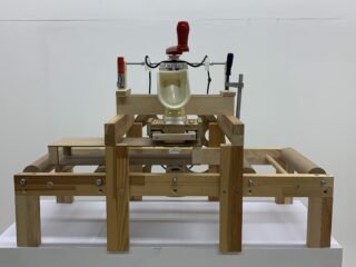 Ein mechanisches Gerät aus Holz mit einem zentralen Kunststoffbehälter, roten und blauen Griffen und Klammern, positioniert auf einer weißen Oberfläche vor einem schlichten Hintergrund.