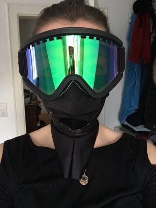 Person mit reflektierender grüner Skibrille und schwarzer Gesichtsbedeckung, an der eine Halskette mit Anhänger sichtbar ist, im Innenbereich.
