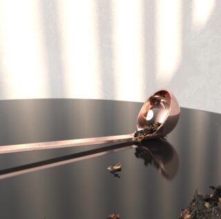 Eine metallische Schöpfkelle mit darin und überall verstreuten Teeblättern liegt unter sanfter Beleuchtung auf einer reflektierenden dunklen Oberfläche.