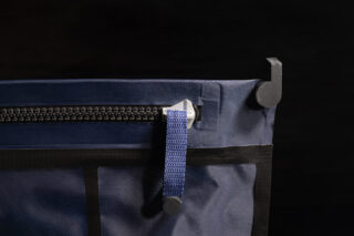 Nahaufnahme einer marineblauen Stofftasche mit schwarzem Reißverschluss, blauem Riemen und Metalldruckknopf, die vor dunklem Hintergrund an einem schwarzen Haken hängt.