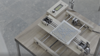 Ein 3D-Drucker steht auf einem Holztisch und druckt kleine graue Objekte auf eine flache quadratische Oberfläche.