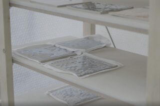 Keramikfliesen mit erhabenen, glänzenden Mustern trocknen auf stoffbezogenen Regalen. Weitere flache, unbearbeitete Fliesen sind auf dem obersten Regal zu sehen.