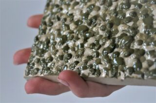 Eine Hand hält eine rechteckige Keramikfliese mit einer strukturierten Oberfläche, die mit glänzenden Unebenheiten in Grün und Creme bedeckt ist.