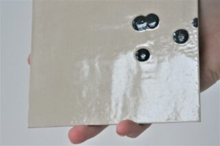 Eine Hand hält eine glänzende, hellbeige Keramikfliese mit mehreren erhabenen schwarzen Punkten auf ihrer Oberfläche.