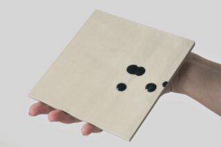 Eine Hand hält vor einem schlichten hellen Hintergrund eine quadratische beige Fliese mit vier unregelmäßig angeordneten schwarzen Punkten auf der Oberfläche.