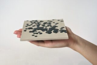 Eine Hand hält eine flache, quadratische Fliese mit unregelmäßigen, erhabenen dunklen Flecken, die über die gesamte Oberfläche verstreut sind.