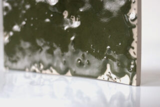 Eine Nahaufnahme einer rechteckigen Keramikfliese mit einer glänzenden, ungleichmäßigen dunkelgrünen Glasur und sichtbaren strukturierten Blasen und Tropfen auf ihrer Oberfläche.