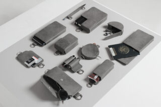 Eine Sammlung grauer Ledertaschen mit Gegenständen wie Schlüsseln, Karten, Münzen, einem Feuerzeug, einer Kamera und einem US-Pass, ordentlich angeordnet auf einer weißen Oberfläche.