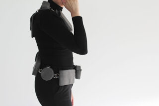 Eine Person in einem schwarzen Rollkragenpullover steht im Profil und trägt einen Werkzeuggürtel und ein Geschirr mit daran befestigten Taschen und Accessoires vor einem schlichten weißen Hintergrund.