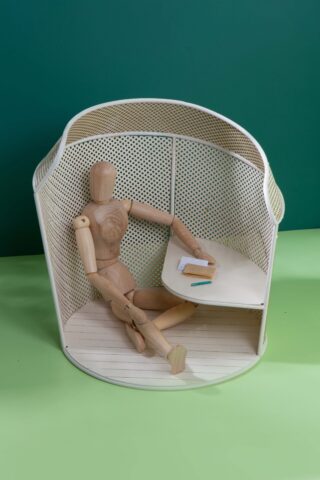 Eine hölzerne Künstlerpuppe sitzt an einem geschwungenen Schreibtisch in einer kleinen perforierten Kabine. Auf dem Tisch liegen ein Notizbuch und ein Bleistift.