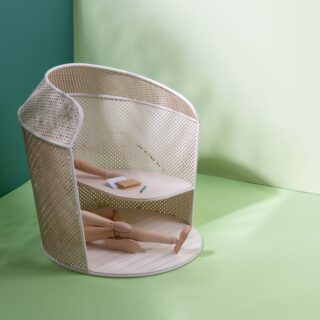 Eine hölzerne Schaufensterpuppe liegt in einem runden, perforierten Gehäuse mit eingebautem Regal und hält einen Bleistift neben einem geschlossenen Notizbuch auf grünem Boden und Hintergrund.