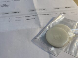 Nahaufnahme von zwei runden Zahnimplantaten in durchsichtigen Plastiktüten, die auf einem ausgedruckten Bestellformular mit Artikelbeschreibungen in deutscher Sprache liegen.