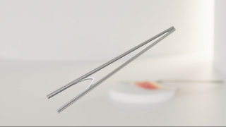 Vor einem unscharfen, hellen Hintergrund sind zwei Essstäbchen aus Metall zu sehen, die in die Luft gehalten werden. In der Ferne sind ein Teller und Essen zu sehen.