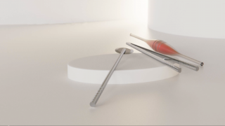 Drei Metallstäbe und ein Metallstab mit roter Spitze ruhen auf einer weißen, ovalen Plattform vor einem minimalistischen, hellen Hintergrund.