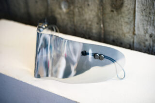 Ein durchsichtiger Gesichtsschutz aus Kunststoff mit einem Metallverschluss und einer elastischen Kordel liegt auf einer weißen Oberfläche, dahinter befindet sich ein Holzhintergrund.