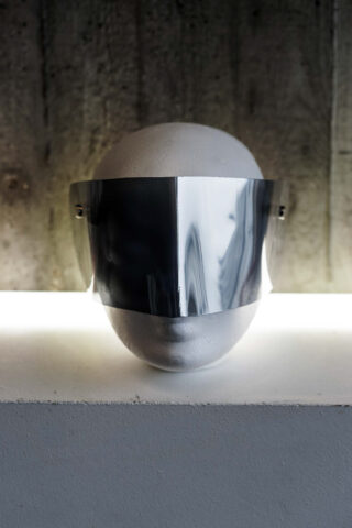 Ein weißer Schaufensterpuppenkopf zeigt ein breites, reflektierendes Metallvisier, das seine Augen und die Oberseite des Gesichts bedeckt. Er steht vor einer Betonwand und wird von hinten beleuchtet.
