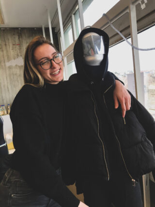 Eine lächelnde Frau mit Brille steht im Haus und hat ihren Arm um eine Person gelegt, die einen schwarzen Kapuzenpullover und eine schwarze Jacke trägt und deren Gesicht von einer reflektierenden Maske verdeckt wird.