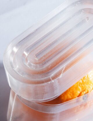 Eine Nahaufnahme eines durchsichtigen Plastikbehälters, der eine Orange teilweise bedeckt, wobei der Behälter eine strukturierte, geriffelte Oberfläche aufweist.